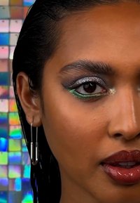 Close-up van een gezicht met zilver en groene oogschaduw, glanzende bruine lippen en zilveren druppeloorbellen. Het haar is nat en de achtergrond heeft iriserende tegels.