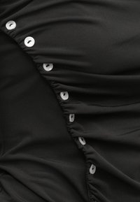 Μαύρο ύφασμα με ρυτιδιασμένη υφή, με λευκά κουμπιά κατά μήκος της μιας πλευράς. Το υλικό φαίνεται λείο και ελαφρώς ελαστικό.