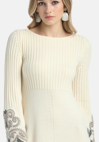 Mlada ženska nosi kremasto rebrasto pleteno obleko z vezenimi sivo vzorci na rokavih in velikimi belimi mramornimi uhani.