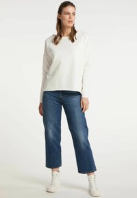 Haut blanc à manches longues avec un col rond, associé à un jean bleu à jambes larges et des baskets montantes blanches. Design simple et épuré.