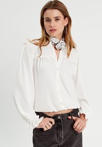Weiße Bluse mit langen Ärmeln aus strukturiertem Stoff, mit Knöpfen vorne, Rüschendetails und elastischen Bündchen, kombiniert mit schwarzen Jeans.