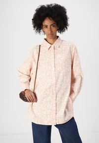 Camisa de botones rosa claro con un patrón floral en naranja. La camisa tiene un corte relajado y mangas largas, combinada con jeans azules.