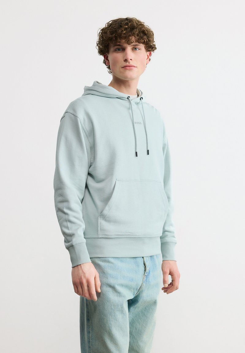 Νεαρός με μπούκλες, φοράει ανοιχτό μπλε φούτερ με κουκούλα και ανοιχτό μπλε τζιν παντελόνι, στέκεται μπροστά σε απλό λευκό φόντο.