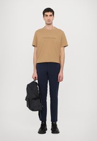Beige cotton t-shirt with embossed logo, navy slim-fit trousers, and a black fabric backpack with adjustable straps. Black sneakers complete the look.