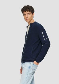 Marineblaue Zip-Up-Jacke aus strukturiertem Material, mit seitlichen Taschen und einer mit Reißverschluss versehenen Armtasche. Getragen über einem hellen Hemd, kombiniert mit hellen Jeans.