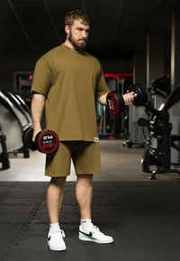 Completo sportivo color khaki, composto da una maglietta a maniche corte e pantaloni corti. Tiene in mano dei manubri neri con accentuazioni rosse. Sneakers bianche con logo nero.