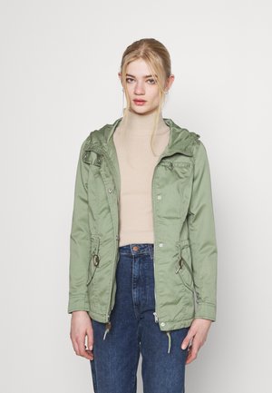 Veste vert olive avec capuche, taille élastique, et fermeture éclair à l'avant, portée sur un haut beige et un jean taille haute bleu. Texture lisse.