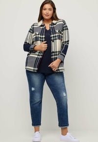 Giacca a camicia a quadri in navy, crema e rosso, caratterizzata da due tasche sul petto, chiusura con bottoni e maniche lunghe. Indossata sopra una maglia navy e jeans.