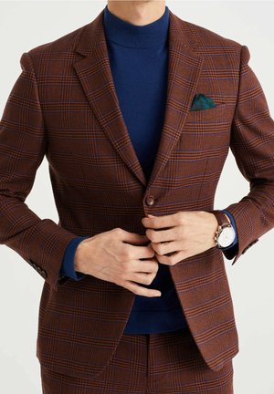 Mężczyzna zapinający brązową marynarkę w kratę na guziki, noszący granatowy golf, zegarek i zieloną poszetkę.