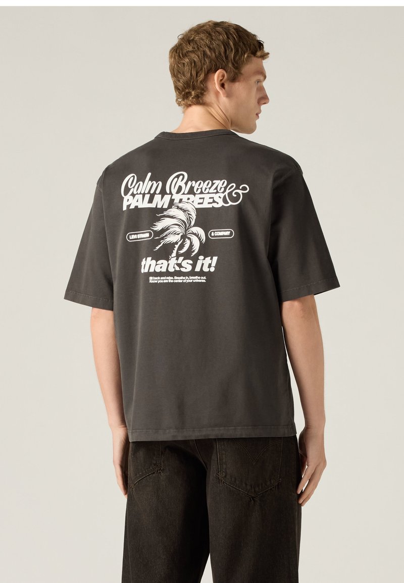Man wearing a black t-shirt with white palm tree graphic and text "Calm Breeze Palm Trees that's it!" on the back, facing sideways.