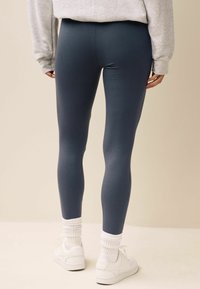 Leggings blu scuro realizzati in un tessuto liscio e elasticizzato, abbinati a calze sportive bianche e scarpe da ginnastica, visti dal retro.