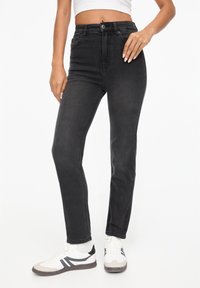Jean en denim noir à taille haute, coupe droite et détails de couture minimaux. Porté avec des baskets blanches avec des accents gris.