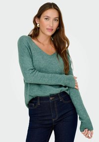 Donna con lunghi capelli castani che indossa orecchini a cerchio d'oro, un maglione verde con scollo a V e jeans blu scuri, in posa con un braccio incrociato.