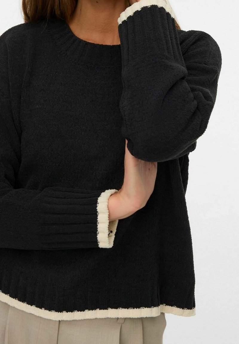 Personne portant un pull texturé noir avec des finitions blanches aux poignets et à l'ourlet, posant avec un bras plié et la main près du visage.
