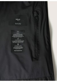Intérieur d'une veste noire montrant l'étiquette MNG Man, une poche zippée, et des étiquettes d'entretien du tissu décrivant des propriétés d'élasticité, de déperlance et de thermorégulation.