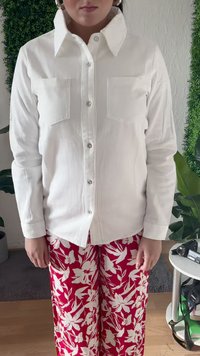 Pessoa vestindo uma camisa branca com botões e colarinho largo, e calças vermelhas com um padrão floral branco, de pé dentro de casa perto de plantas e uma mesa.