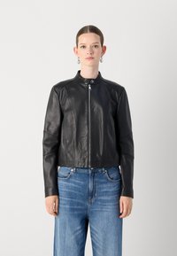 Črna usnjena skrajšana jakna z visokim ovratnikom in zadrgo. Ima teksturirane panele in dolge rokave, kombinirana z svetlo modrimi džins hlačami.