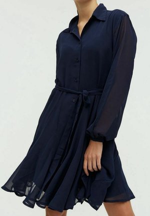 Vrouw die een marineblauwe jurk met knopen draagt, met lange doorschijnende mouwen en een ceintuur in de taille, terwijl ze de zoom van de jurk vasthoudt.