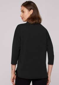 Top negro de manga tres cuartos con cuello redondo y aberturas laterales, hecho de tela suave. Presenta un corte holgado y un dobladillo curvado.