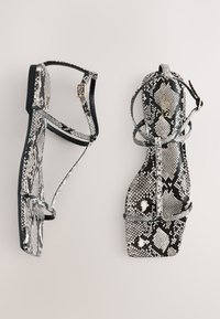 Juodos ir baltos gyvatės rašto šlepetės su plokščiu padu, turinčios kryžminius dirželius ir struktūrizuotą pakulnę. Lygus paviršius, reguliuojamas sagtis.