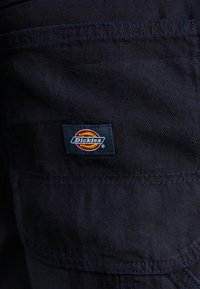 Black fabric features with a Dickies brand tag in blue, red, and yellow. The tag is rectangular with an embroidered logo and white text.