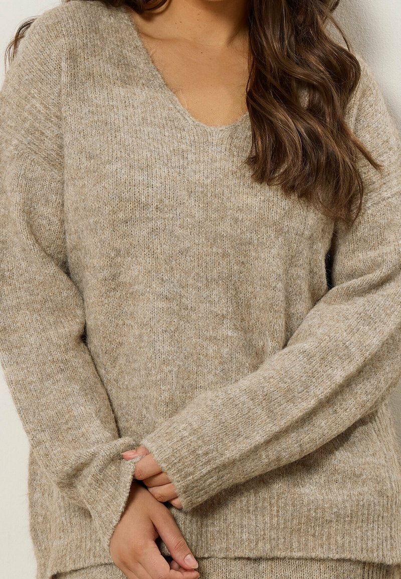 Gros plan d'une personne portant un pull en maille beige doux à manches longues et col en V, les mains doucement jointes à la taille.
