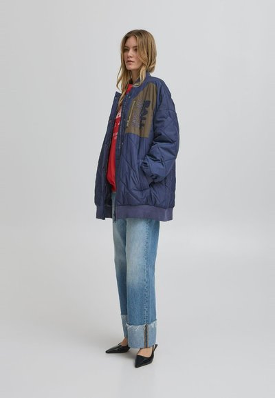 Marineblå quiltet jakke med olivengrønne detaljer, med et grafisk print. Båret over en rød skjorte og parret med lyseblå jeans.