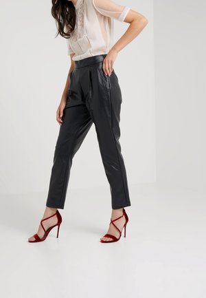 Vrouw die een doorschijnende witte blouse draagt, hoge zwarte leren broek en rode sandalen met hoge hakken en bandjes, staande op een witte vloer.