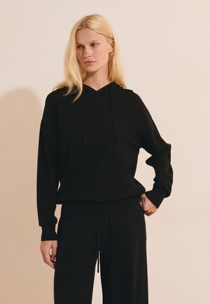 Mujer rubia que lleva una sudadera con capucha negra y pantalones negros a juego, de pie frente a un fondo beige liso.