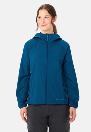 Vrouw die een blauw vest met rits en capuchon draagt en donkere broek, kijkt iets opzij met een neutrale achtergrond.