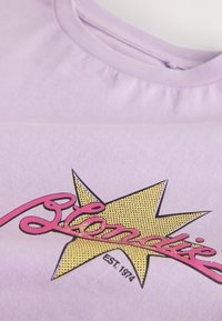 T-shirt lilas avec une étoile jaune vintage et un texte cursif rose « Lémandie », incluant « EST. 1974 » imprimé sous l'étoile.