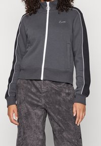 Nike mikina na zip v tmavě šedé barvě, s bílými akcenty a vysokým límcem. Vyrobena z měkkého materiálu s přiléhavým střihem a bočními kapsami.