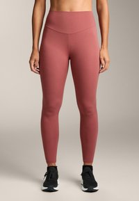 Magasan dekoltált, poros rózsaszín leggings, rugalmas anyagból készült, sima felülettel és teljes hosszúságú kivitelben, fekete sportcipővel párosítva.