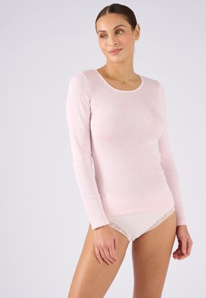 Donna con capelli castani con top rosa a maniche lunghe e biancheria intima bianca con bordi in pizzo, in piedi davanti a uno sfondo chiaro uniforme.