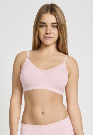 Frau trägt hellrosa Spitze-BH und passende Unterwäsche, mit glattem langen Haar, blickt nach vorne vor einem einfarbigen Hintergrund.