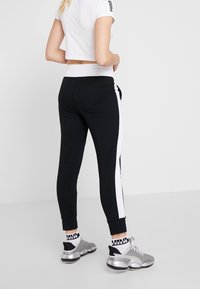 Svarta joggers med vita sidostråk, åtsittande design, ankelfållar, bärs med silverfärgade sneakers och mönstrade strumpor, visade från baksidan.