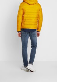 Gul pufferjakke med hætte, blåt lagdelt skjorte, jeans med medium vask og hvide sneakers, set fra ryggen. Tekstureret, quiltet design.