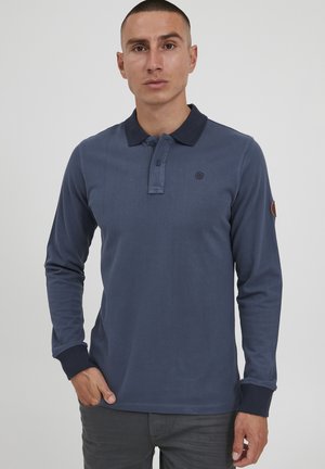 Tamno plava dugosleeve polo majica s tamnim ovratnikom, otvorom za tri dugmeta i malim logotipom na grudima. Ima rebraste manžete i glatku teksturu.