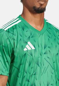 Grünes Sporttrikot mit einem strukturierten Muster aus dreieckigen Formen. Verfügt über einen V-Ausschnitt und drei weiße Streifen auf den Schultern.