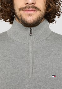 Pulover gri cu fermoar, cu guler striat, având un fermoar până la jumătate și o mică etichetă cu logo în colțul din jos. Textură tricotată moale.