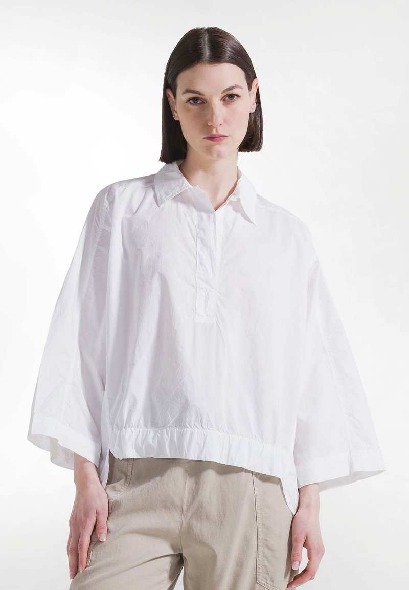 Femme aux cheveux lisses et foncés portant un chemisier blanc ample à col et un pantalon beige clair, debout devant un fond blanc uni.