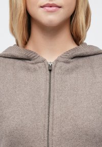 Barna, cipzáros kapucnis pulóver texturált kötött anyagból, kapucnival és ezüst cipzárral. A fazon bordázott díszítéseket tartalmaz a nyak körül.