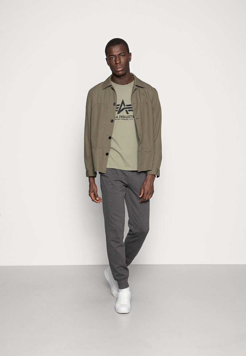 Chaqueta ligera verde oliva sobre una camiseta verde clara con un logo, combinada con joggers gris oscuro y zapatillas blancas.