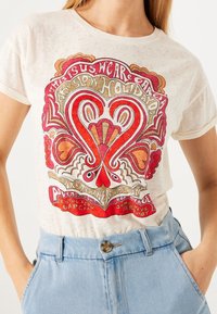 Camiseta de algodón color crema con un gran diseño de corazón rojo y dorado, con intrincados patrones en espiral y texto. Mangas cortas y corte relajado.