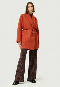 Abrigo de mezcla de lana en color óxido con cinturón, mangas anchas y dos bolsillos frontales, combinado con pantalones acampanados estampados en azul marino y naranja.