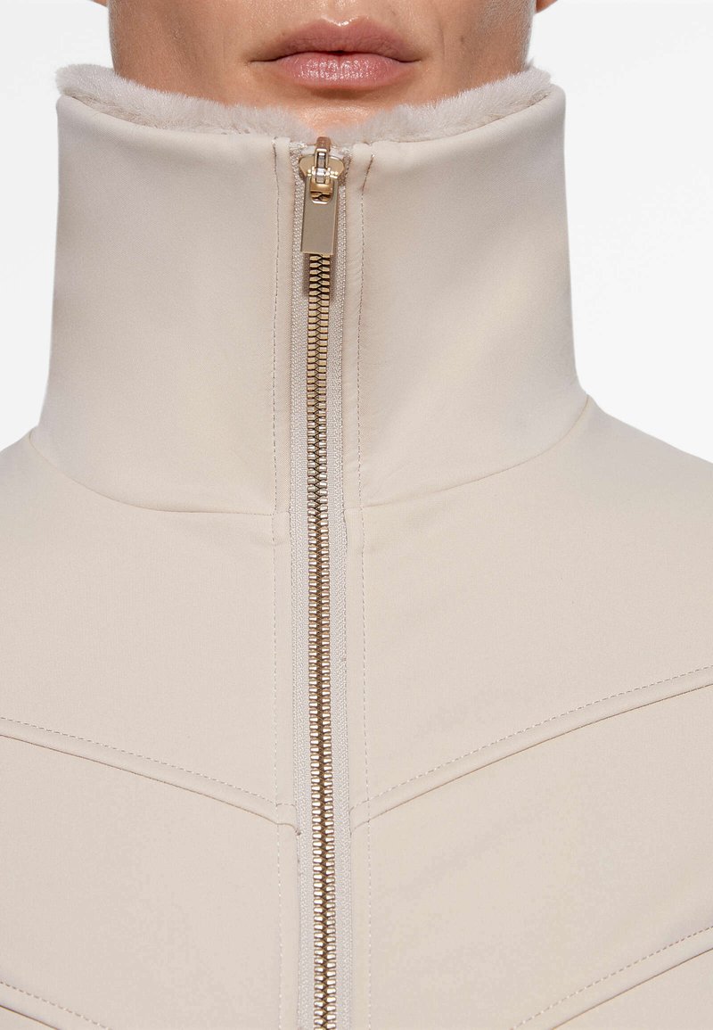 Chaqueta beige de cuello alto con cierre de cremallera, que cuenta con un suave forro de piel, textura lisa y detalles de costura en diagonal para un aspecto estructurado.