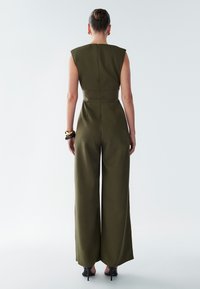 Olivově zelený overal se širokými nohavicemi, strukturovaným designem ramen, přiléhavým pasem, zapínáním na zip vzadu a nošený s černými sandály na podpatku.