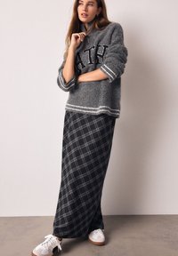 Pull gris flou avec les lettres "ATH" et des rayures blanches ; associé à une longue jupe plaid noire. Chaussures : baskets blanches avec semelles marron.