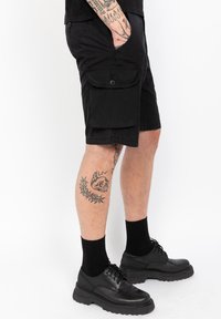 Zwarte cargoshorts met zijzakken, gemaakt van lichtgewicht materiaal. Draagt zwarte sokken en schoenen, en heeft een tatoeage op het been.