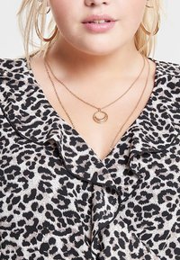 Top med leopardtryck och volangkrage i svart. Guldhalsband med flera lager och en cirkulär hängsmycke. Slät tygtextur.